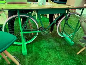 A bicycle frame is incorporated into the base of a table. The seating area is covered with green artificial grass.
