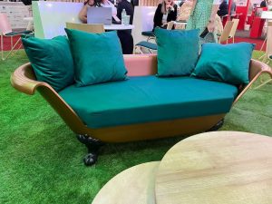 A gold and green cushioned sofa shaped like a bathtub with wooden armrests is placed on artificial grass. 