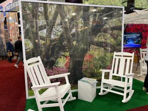 Two white rocking chairs face a backdrop of a lush garden scene with trees and flowers. A small table between the chairs holds a book.