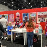 Authors sign books at a booth with bright posters and book displays. People line up while staff assist and interact.