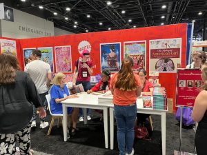 Authors sign books at a booth with bright posters and book displays. People line up while staff assist and interact.