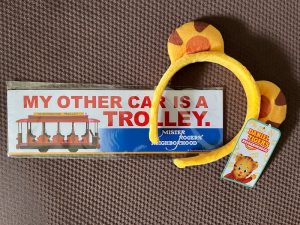 A bumper sticker reads "My Other Car Is a Trolley" with a trolley image, and a yellow tiger ear headband with a Daniel Tiger's Neighborhood tag next to it.