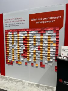 A display board covered with colorful sticky notes spells out "HERO" under signs about librarians' superpowers and community impact. A small table with buttons is visible on the right.